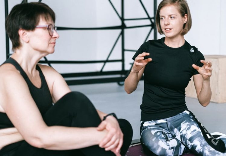Two women in sportswear sitting on a yoga mat, having a detailed discussion in a gym.