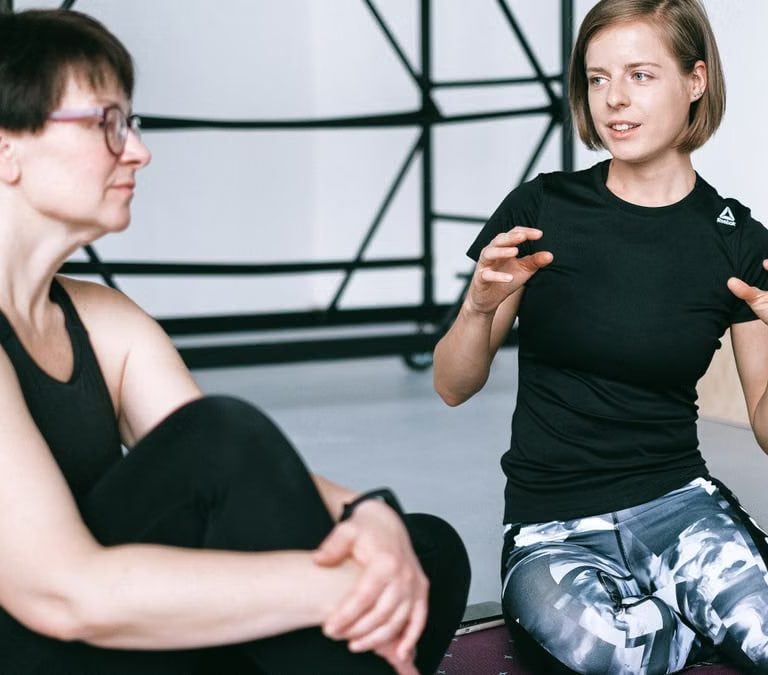 Two women in sportswear sitting on a yoga mat, having a detailed discussion in a gym.